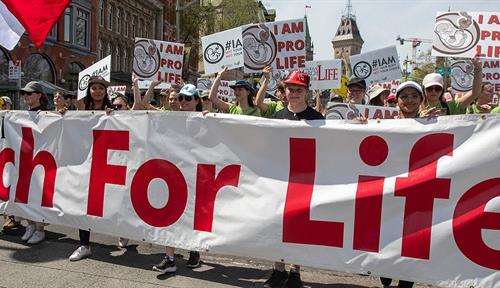 People holding placards march behind a large white banner with “National March for Life” written on it in red. There are buildings in the background.