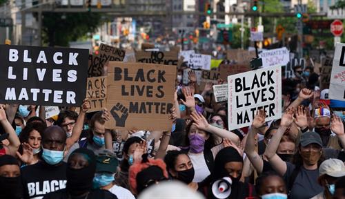 Photograph of a crowd of mask-wearing protestors in a city street holding banners that read “Black lives matter” and one that reads “Defund the police.”