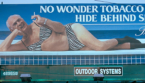 A billboard depicts an older male tobacco executive lounging in a bikini while smoking a cigarette. 