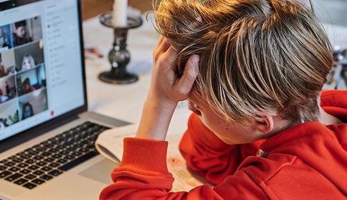 A young boy in a red-orange sweatshirt sits at a table in front of a laptop computer with his head in his hands. Other participants in a Zoom class are visible on the screen.