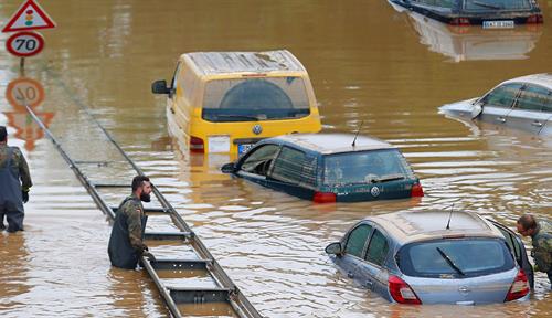 Several cars are partially submerged in a road flooded with waist-deep water; some people wade among the cars.