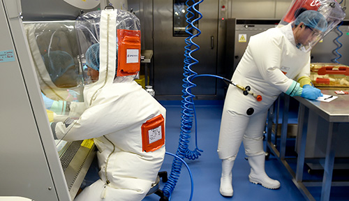 Lab researchers wearing full protective suits, supplied with clean air, are shown in a lab.