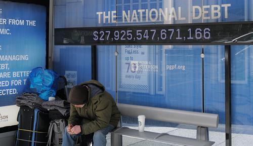 Photo shows an unhoused man with his belongings resting in a covered bus stop in Washington, DC, in February 2021. A digital sign in the bus stop tracks upticks in the US national debt.