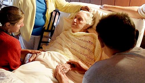 Photograph of an elderly woman lying in bed. Standing and sitting around her are family members, two of them holding her hands. A hospice aide and a bottle of lotion are seen at the foot of the bed.