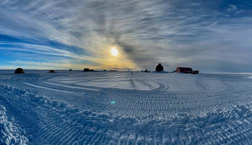 Photograph of a flat, desolate, ice- and snow-covered landscape with the Sun low in the sky. A few structures are in the background.