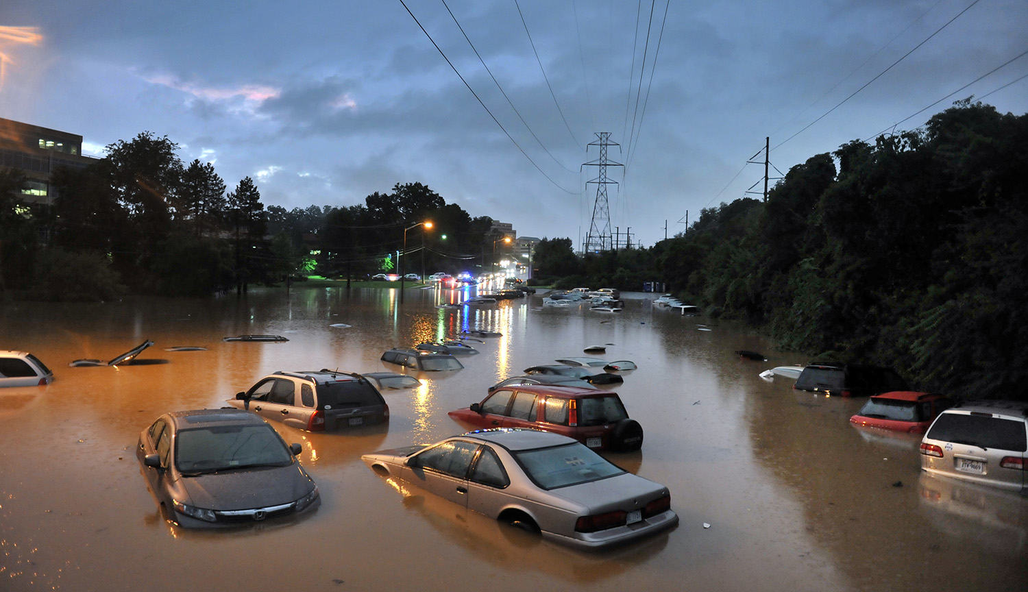 Photograph of a wide flooded parking lot with more than a dozen marooned, partially submerged cars. Dark storm clouds loom overhead.