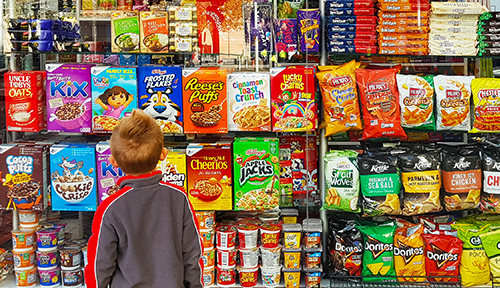 Photo of a young boy standing in front of a supermarket shelf filled with colorful bags of chips, boxes of cereals and other processed snack products.