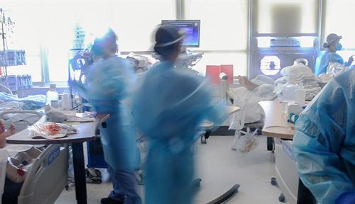 Photograph of four health workers clad in blue gowns, face shields and masks. They are moving from bed to bed in a hospital room. Three beds are visible, as well as various kinds of medical equipment including ventilators and drips.