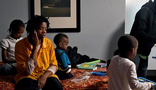 A family of four gathered in a hotel room, the father standing up by a small table figuring out food, the mother and children sitting on the bed, which has toys and books on it.. 