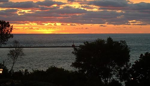 Photograph of a lakeshore fringed with trees as the sun sets, with lights gleaming out from a few houses.