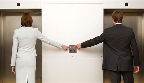 Photograph shows a back-view of a man and woman in business attire standing in front of two elevators and reaching for the buttons.