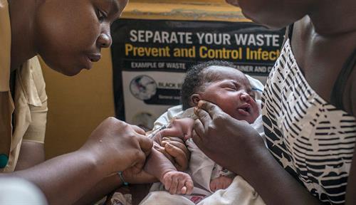 Two women sit facing each other. The woman on the right is holding a baby, supporting the baby’s head with her hand. The woman on the left is administering vaccine into the baby’s arm.