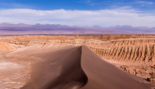 A massive brownish sand dune, steep orange-brown rocks behind it and purplish mountains in the distance