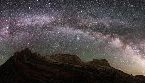 Photograph at night, with mountainous terrain and desert floor. In the sky one can see thousands of stars and the disk of the Milky Way galaxy.