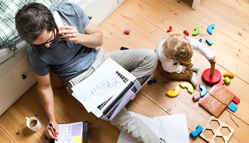 Photo shows a dad seated on the floor with a laptop balanced on his lap and writing in a planning calendar while talking on the phone. A baby sits at his knee, playing with blocks.