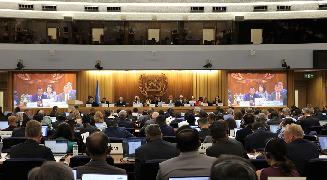 Photograph of a meeting taking place in a large room, with panelists up on stage and a packed audience watching and takes notes on laptops. The IMO logo is up on the wall behind the stage.