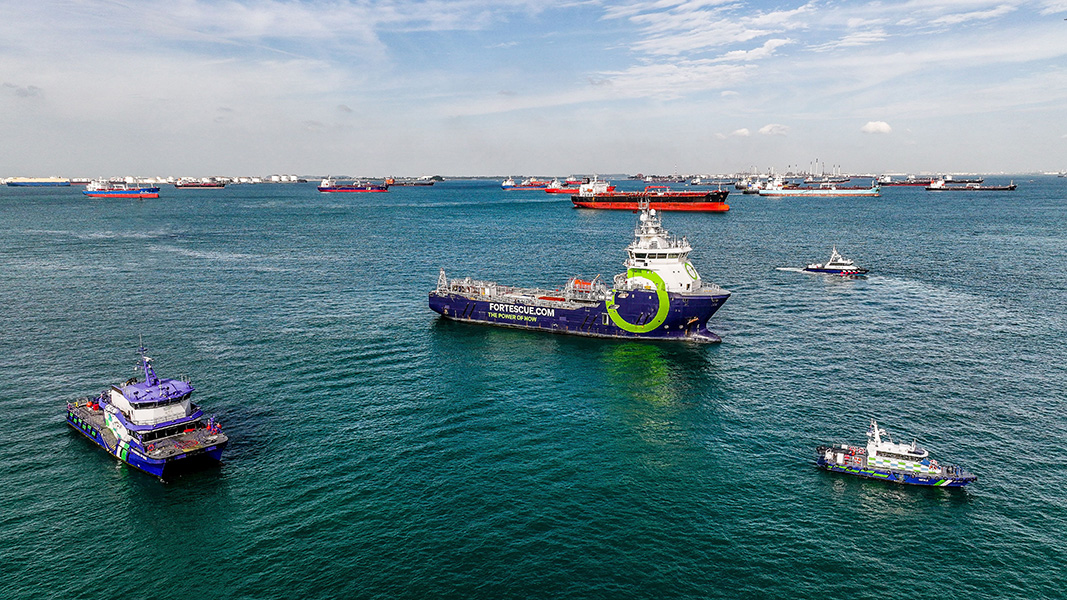 Photograph a variety of vessels moored in a seaport. Near the front is a ship that says “fortescue.com, the power of now” on its side.