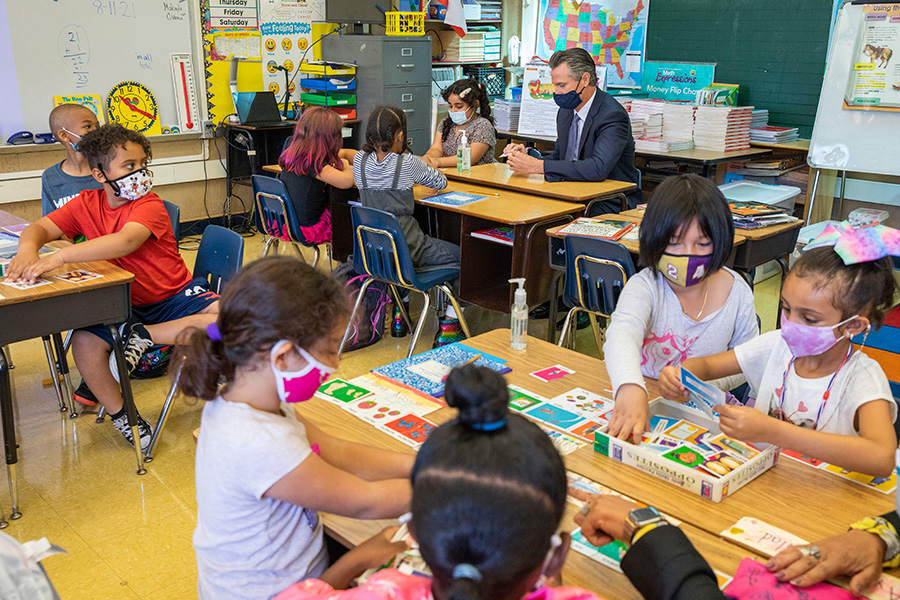 Governor Gavin Newsom Photograph of an elementary school classroom. Children wearing colorful masks are sitting at tables. Governor Gavin Newsom, wearing a suit and dark mask, is sitting at one of the tables.