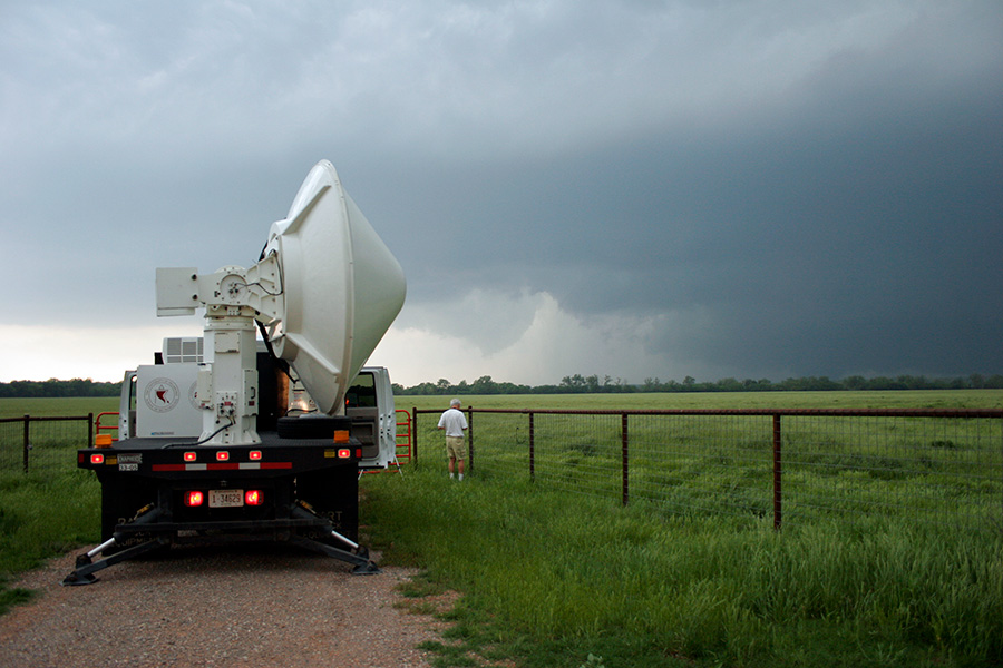 Mobile radar A truck seen from the back with a giant radar contraption in its bed, a person stands nearby looking at tall clouds spawning a tornado in the distance