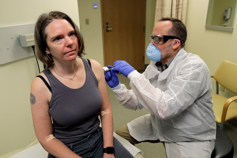 Photograph of a woman in a tank top sitting on a bench receiving a shot in the arm. A man wearing a mask, goggles, gloves and a white coat is administering the shot.