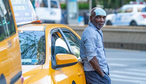 A man stands outside a taxicab, face mask on his forehead, looking at the camera