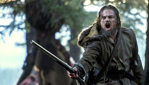 Still photograph from the 2015 movie The Revenant. Frontiersman Hugh Glass (Leonardo DiCaprio) is brandishing a rifle and shouting in fury, beneath trees.