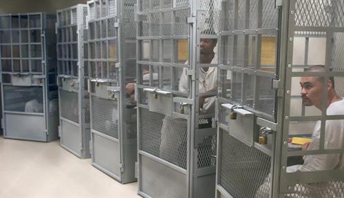 Photo shows multiple, booth-sized metal cages in a room together, each with a prisoner inside. They are able to talk and see each other but cannot move freely. 