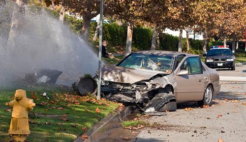 Photograph of a smashed-up car that ran off the road into a lamppost. A fire truck and police cars are in the background.