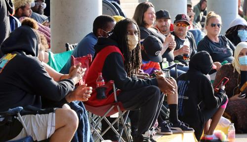 Photo shows a section of a long queue of people waiting for help with unemployment insurance claims outside of an office. Some people are sitting in folding chairs.