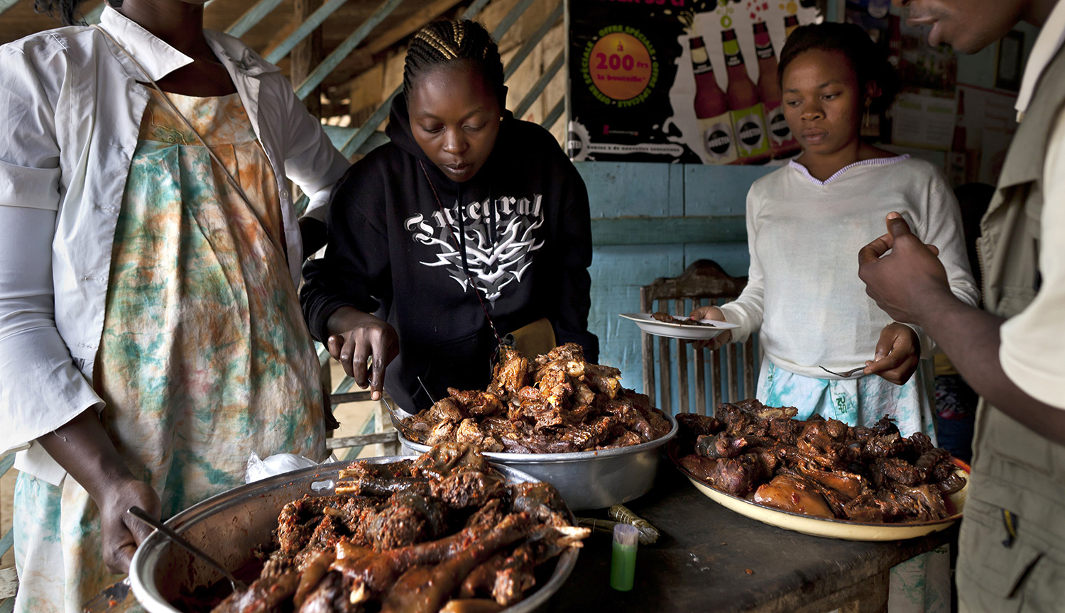 Large platters hold monkey meat that has been cooked. Several women stand behind the platters.