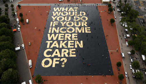 Aerial photograph of a large city square that contains a huge poster with the words “What would you do if your income were taken care of?”